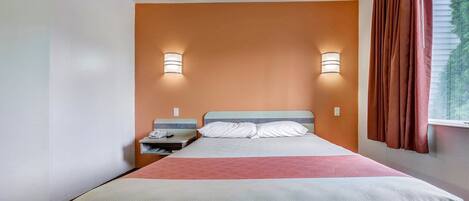 Una habitación de hotel con cama, mesita de noche, lámpara y ventana con cortinas.