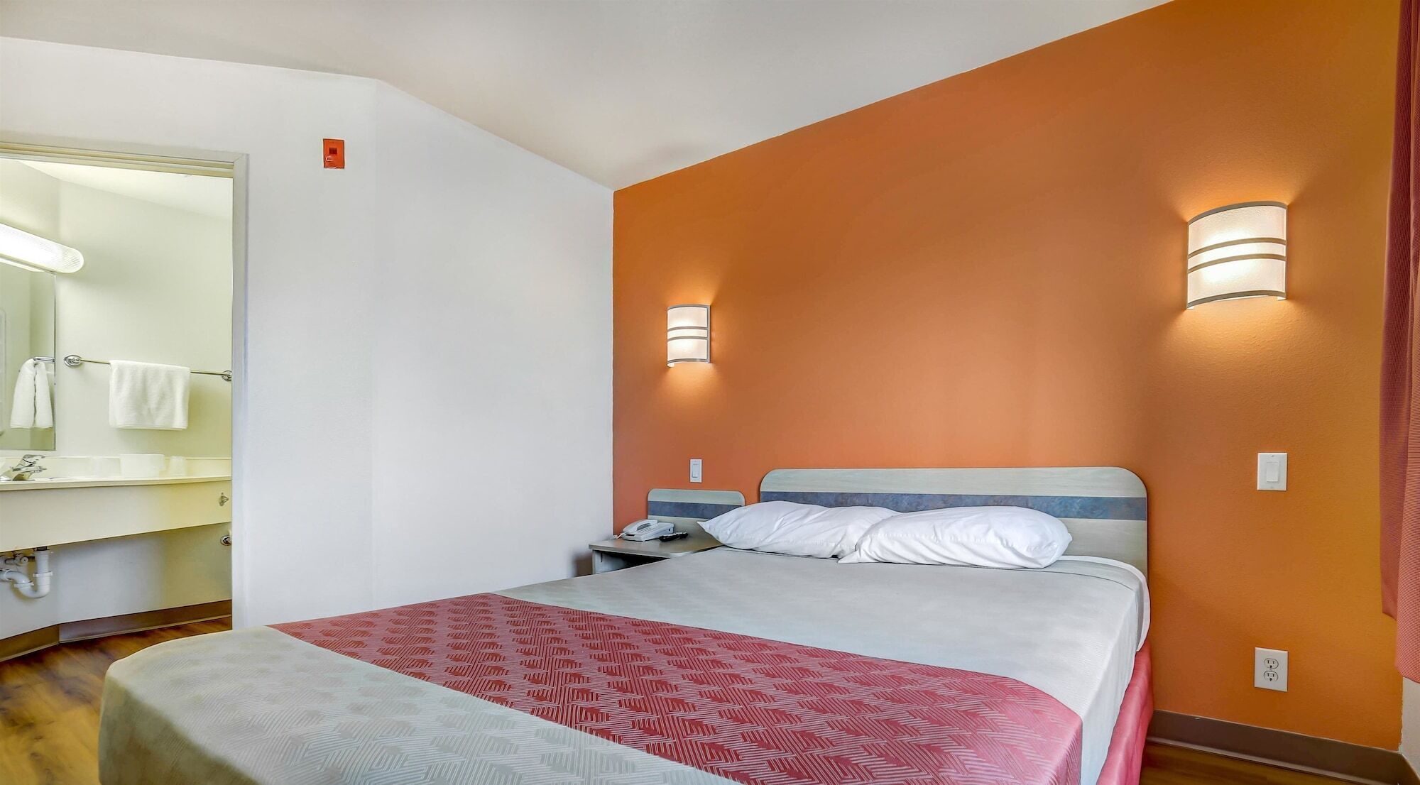 Habitación de hotel con cama, mesita de noche, teléfono, espejo y baño visible a través de una puerta abierta.