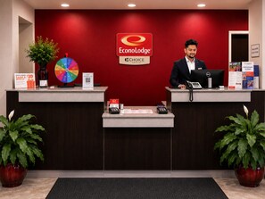 A hotel reception desk with a red wall, a logo for EconoLodge, and a sign for Choice Hotels. There are two receptionists, a plant, and a wheel of rewards. - Econo Lodge Menomonie (Menomonie)