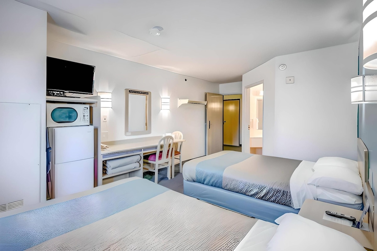 A hotel room with two beds, a desk with a chair, a TV, a microwave, and a door leading to another room.