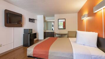 Una habitación de hotel con una cama, un escritorio con computadora, televisor y una kitchenette pequeña.