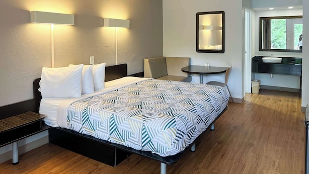 A hotel room with a bed, bedside table, chair, small round table, and a bathroom with a sink and mirror.