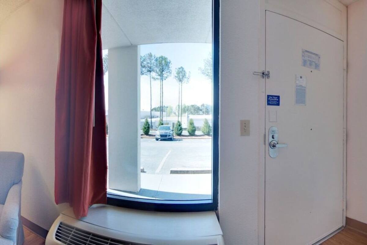 A hotel room with a bed, a chair, a desk, a TV, and a window with a view of trees and a parking lot.