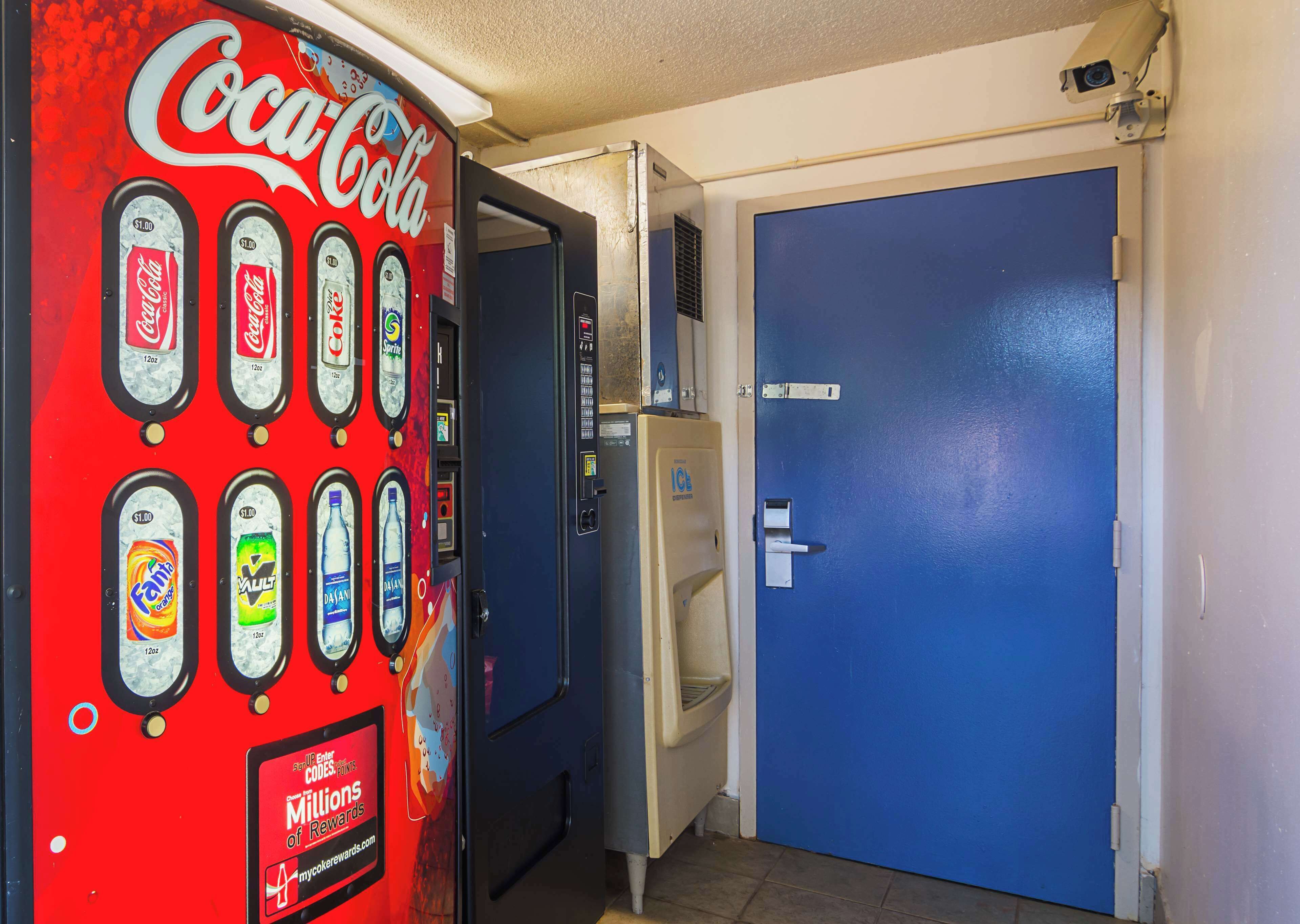 A vending machine with various drink options and a blue door with a security camera above it.