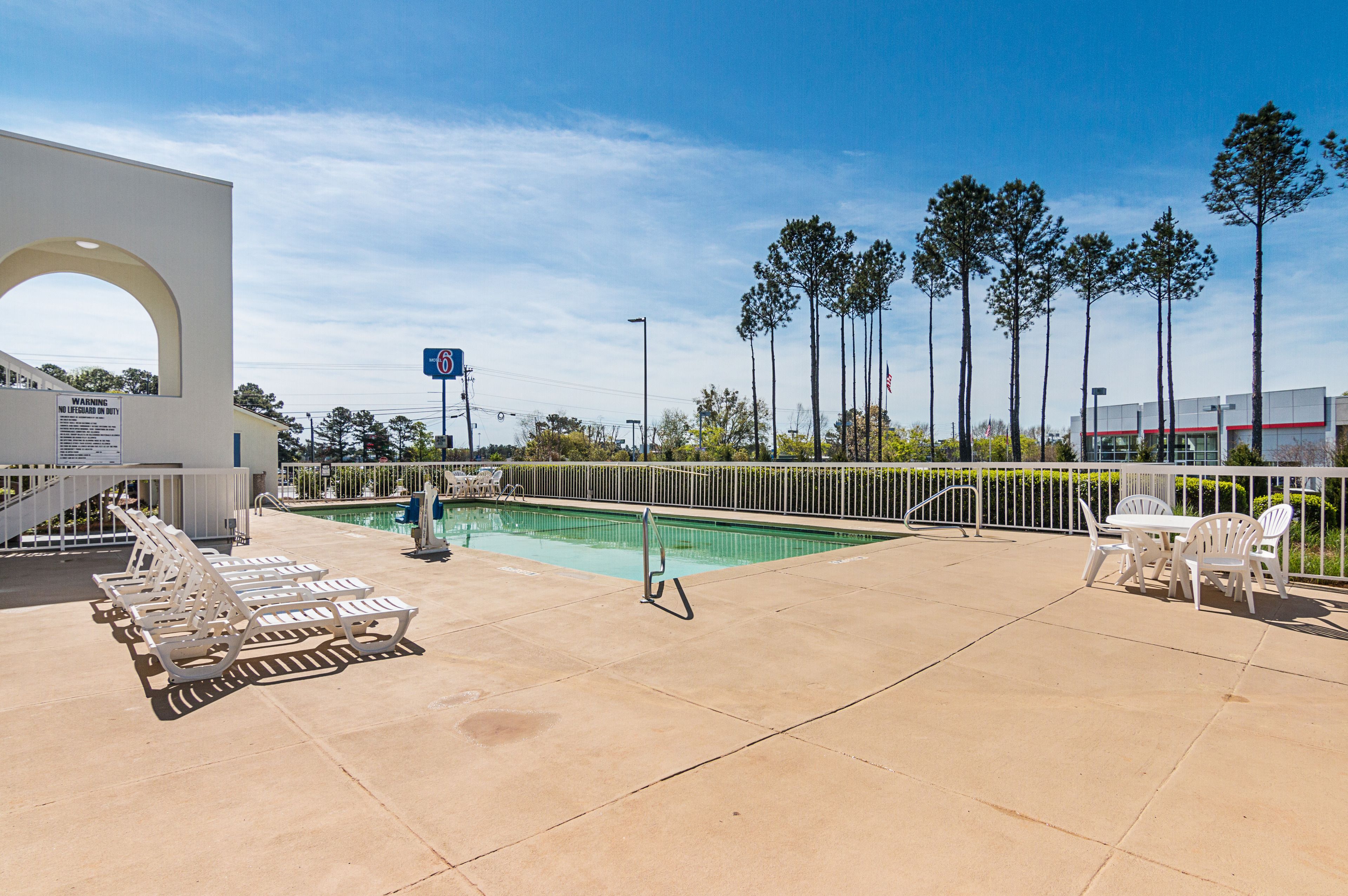 A pool area with lounge chairs and a table, surrounded by a fence and tall trees.