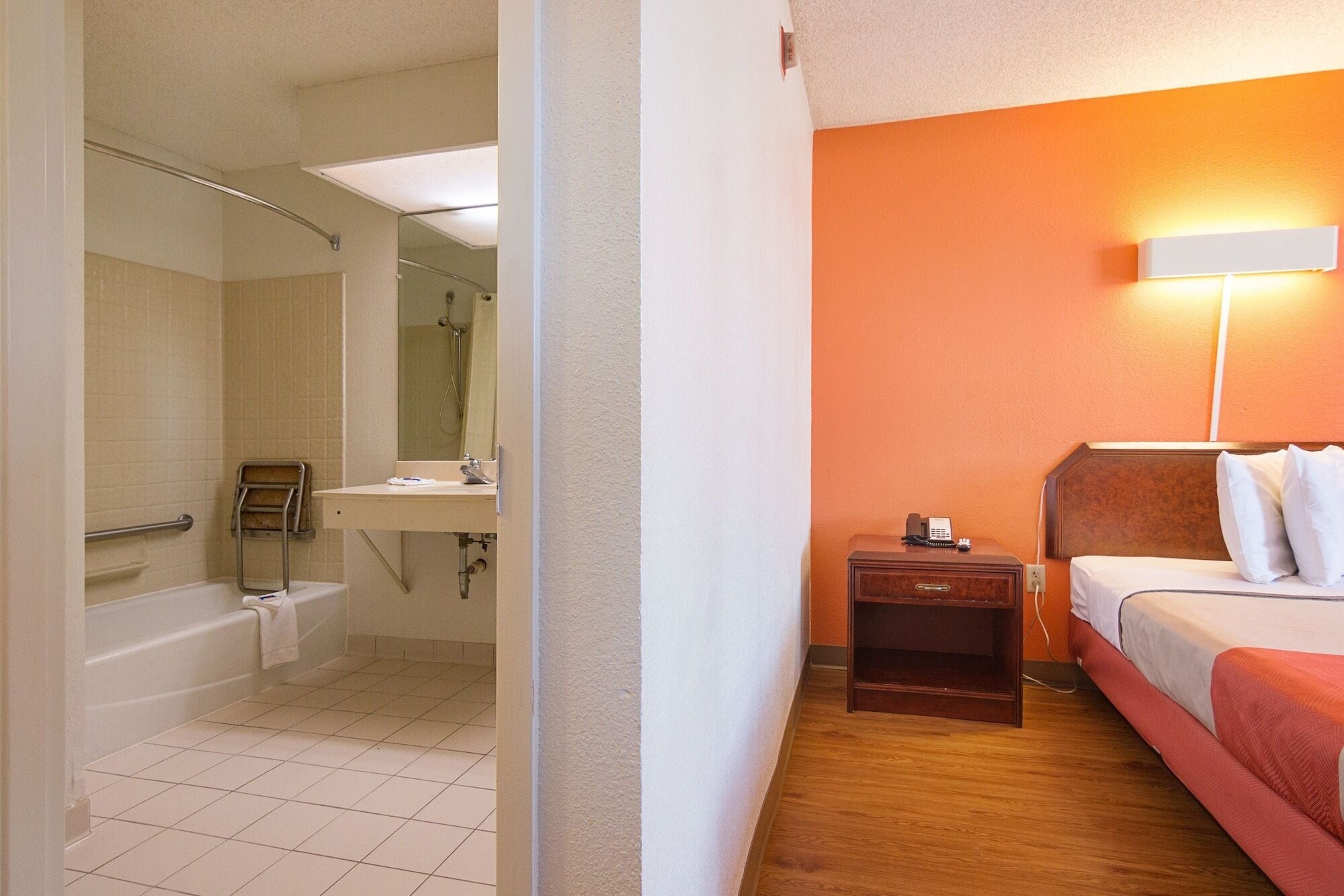 A hotel room with a shower, a sink, a bedside table with a lamp, and a bed.