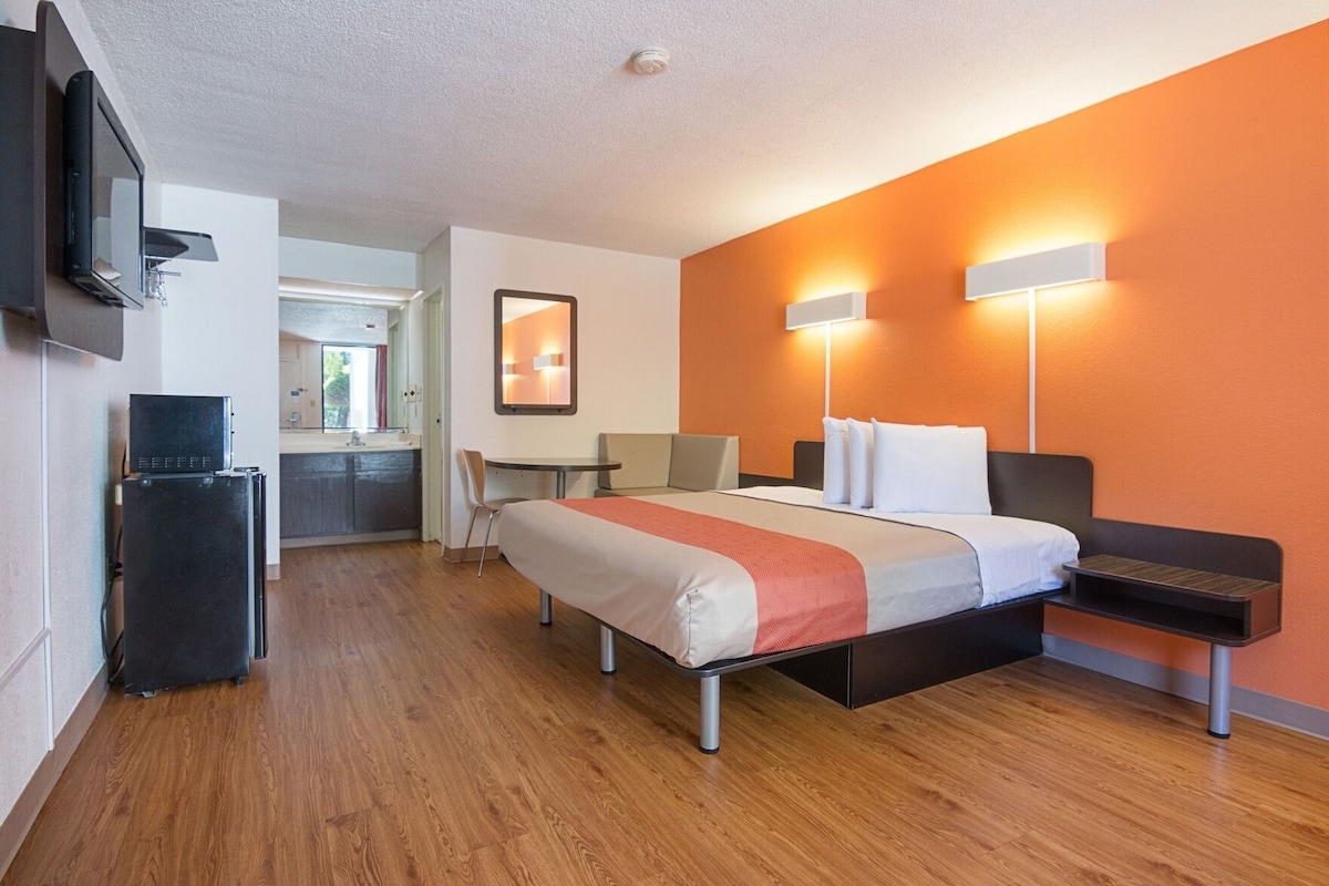 A hotel room with a bed, bedside tables, a desk, a chair, a refrigerator, and a TV mounted on the wall.
