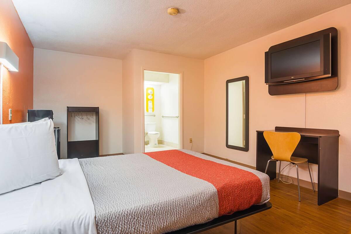 A hotel room with a bed, a desk, a chair, a TV, and a bathroom visible through an open door.