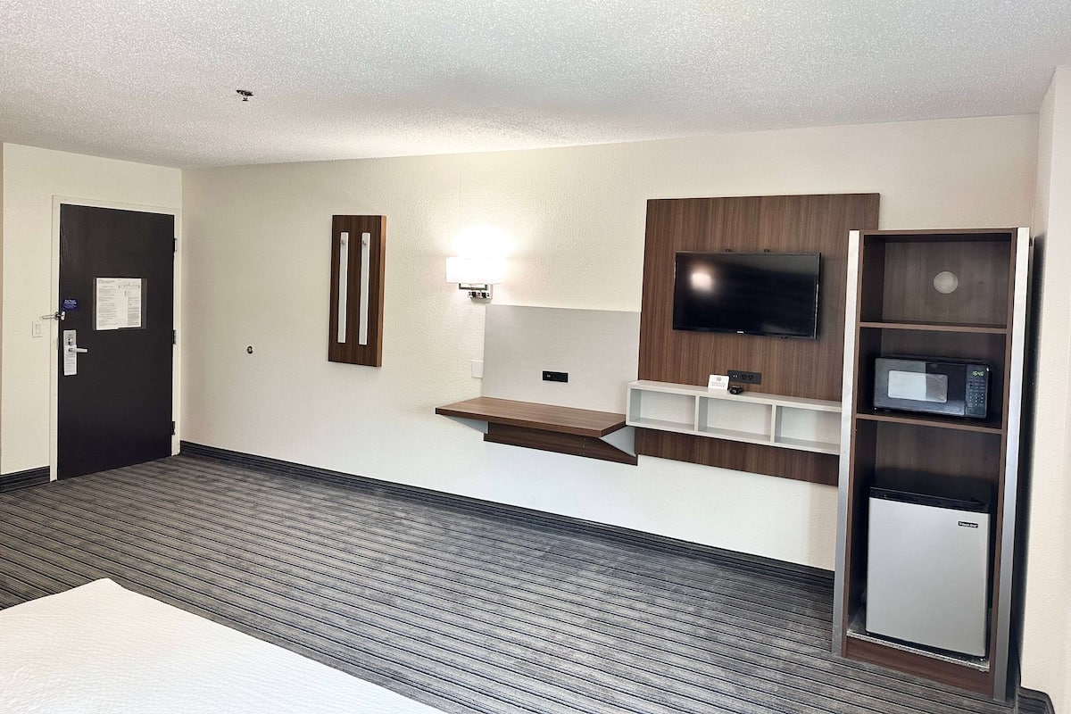 A hotel room with a bed, a flat-screen TV mounted on the wall, a microwave, and a refrigerator built into the shelving unit.