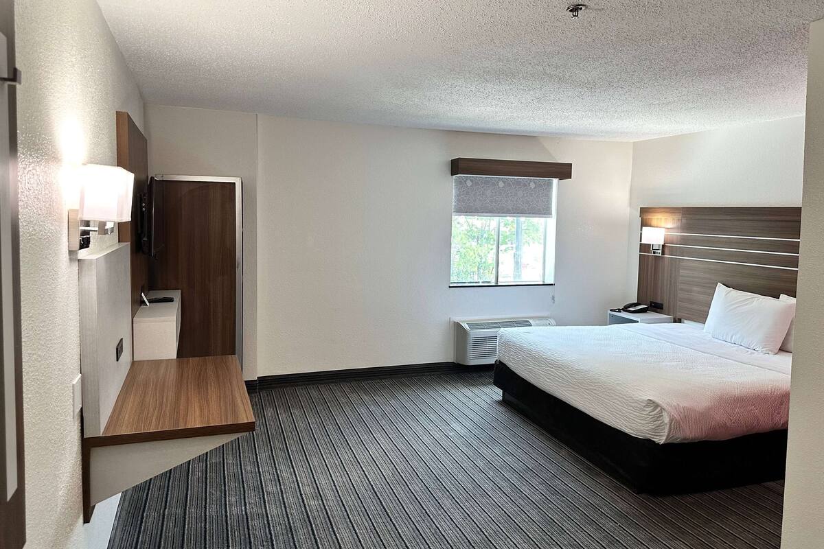 A hotel room with a bed, bedside table, lamp, and a small desk with a chair.