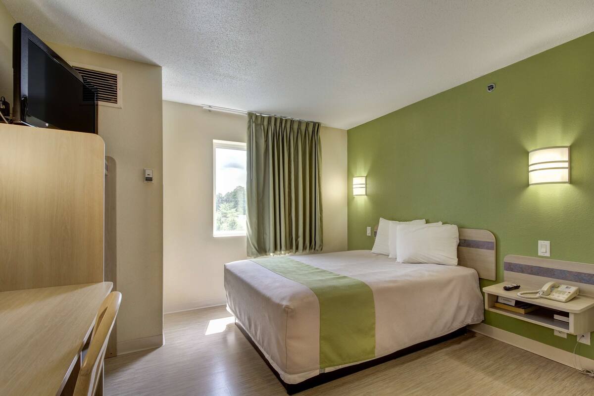 Ein Hotelzimmer mit Bett, Nachttisch, Lampe und einem Fenster mit Vorhängen.