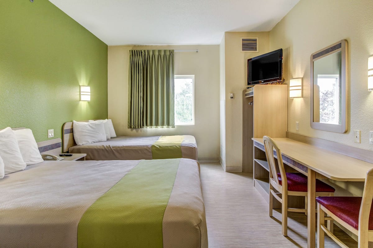 Ein Hotelzimmer mit zwei Betten, einem Schreibtisch mit Stuhl, einem Fernseher und einem Fenster mit Vorhängen.