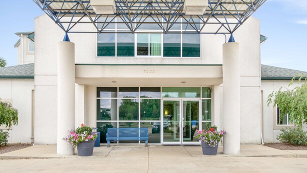 A building entrance with a blue bench, two potted plants, and a clear glass door. - Motel 6 Medina, OH - Cleveland (Medina)