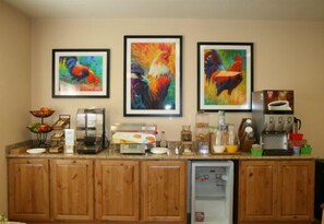 A kitchen area with wooden cabinets, a refrigerator, a coffee maker, and three framed paintings of roosters.