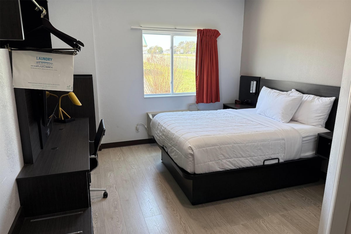 Habitación de hotel con cama, escritorio, silla, armario y ventana con vistas a árboles.