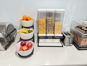 Una encimera de cocina con un bol de fruta, un dispensador de cereales, un tostador y un paquete de café.