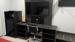 A compact office space with a flat-screen TV, a small refrigerator, a yellow desk lamp, and a black office chair.