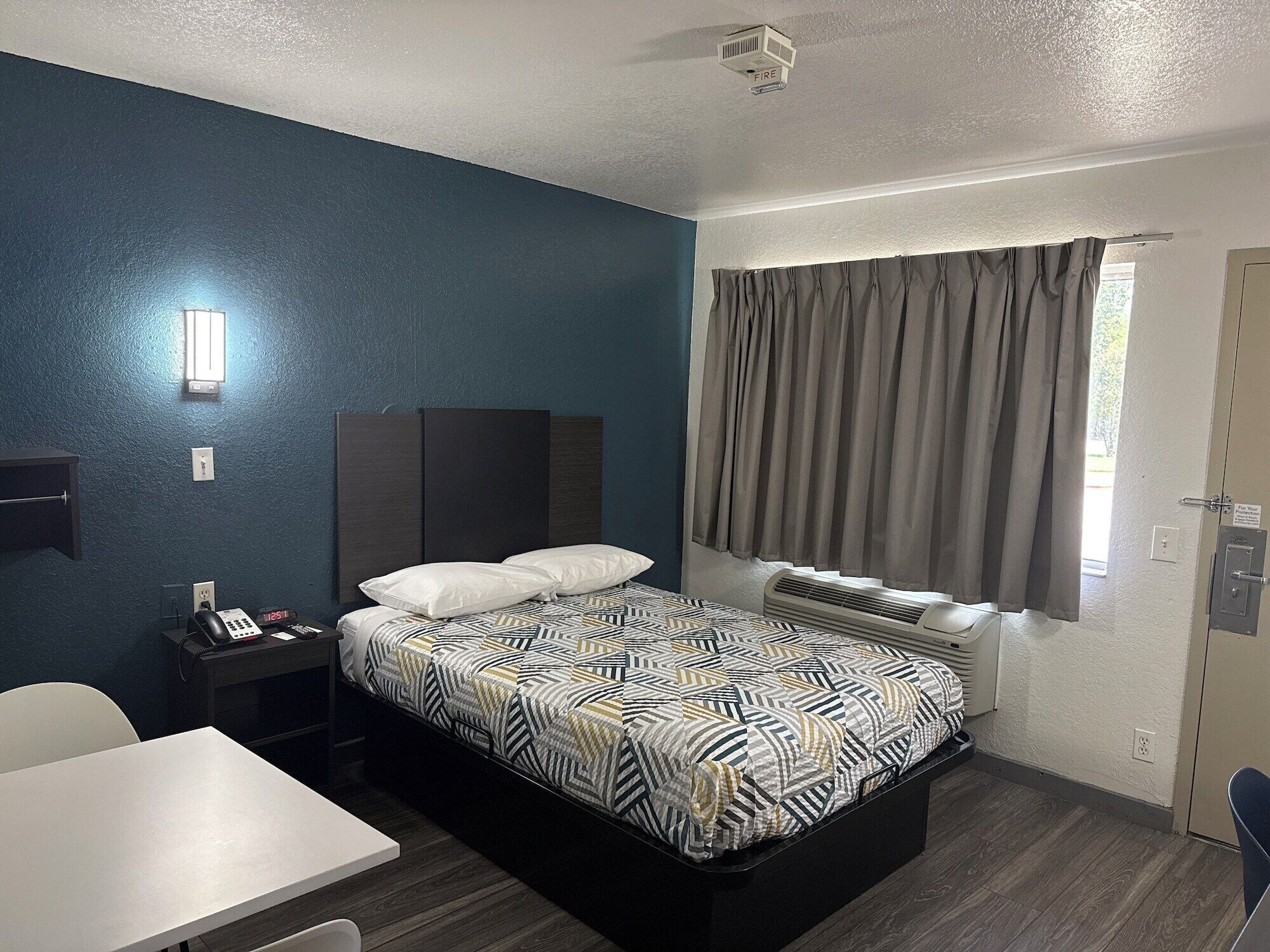 A hotel room with a bed, a desk, a chair, a window with curtains, a wall-mounted heater, and a door with a sign.