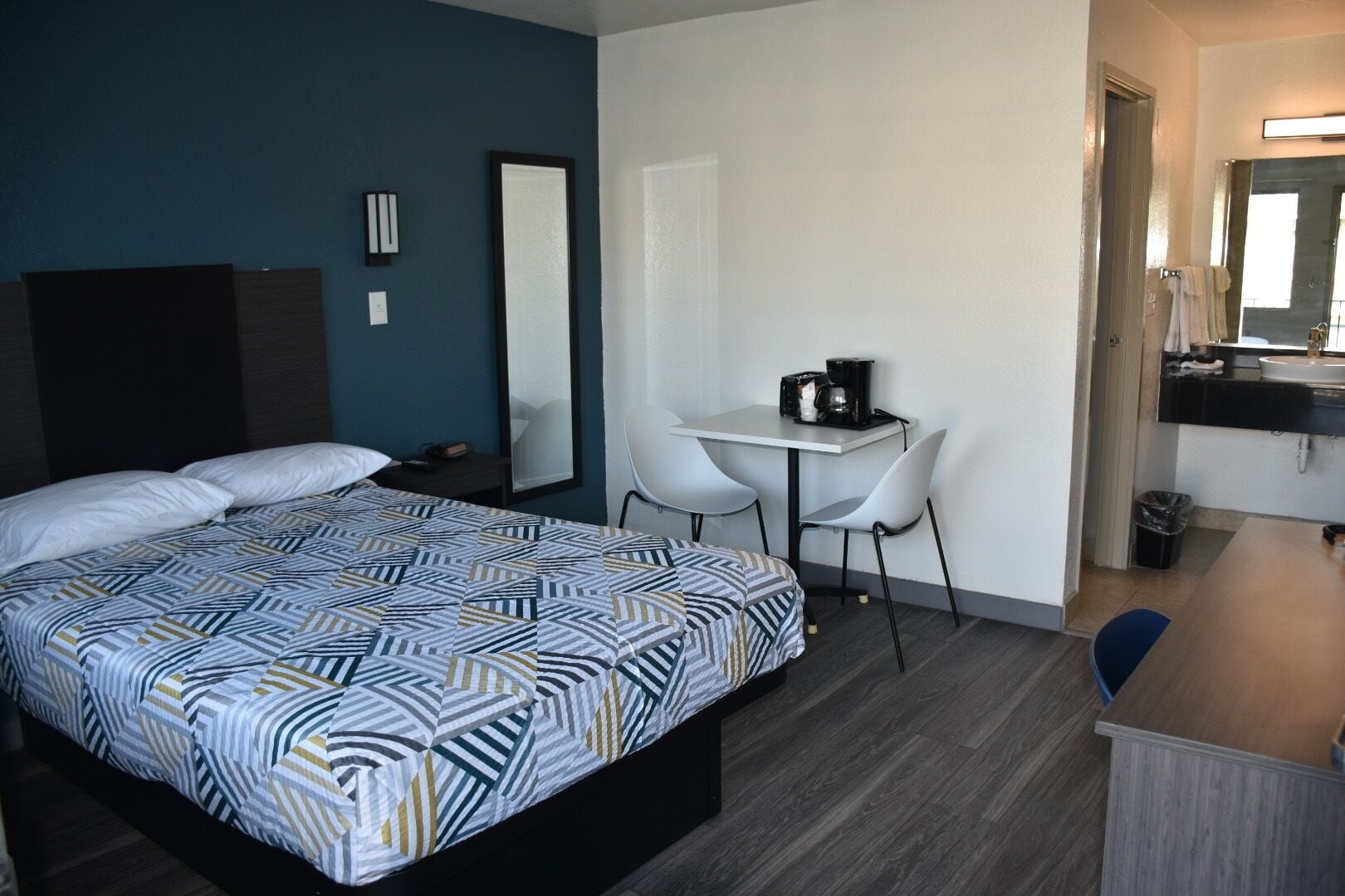 Habitación de hotel con cama, escritorio, dos sillas, una mesita con cafetera, cubo de basura y espejo.