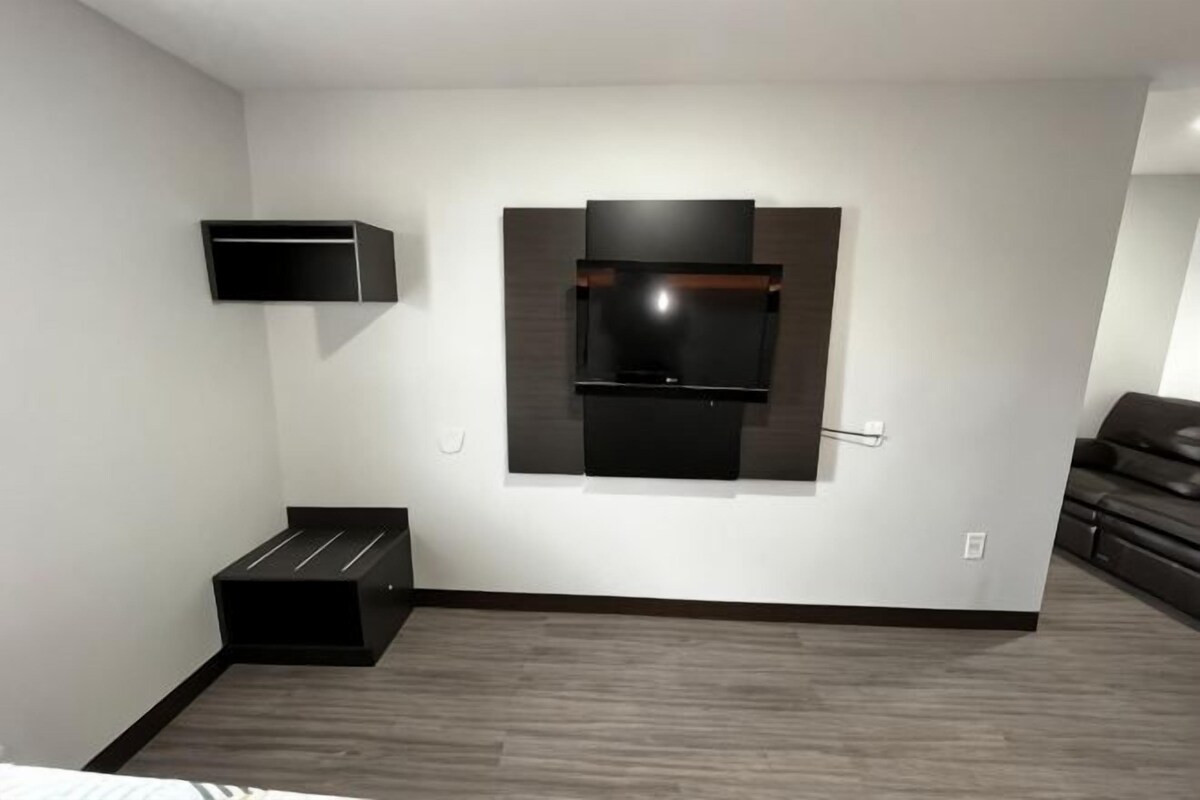 A modern hotel room with a flat-screen TV mounted on the wall, a small black storage unit, and a dark-colored sofa.