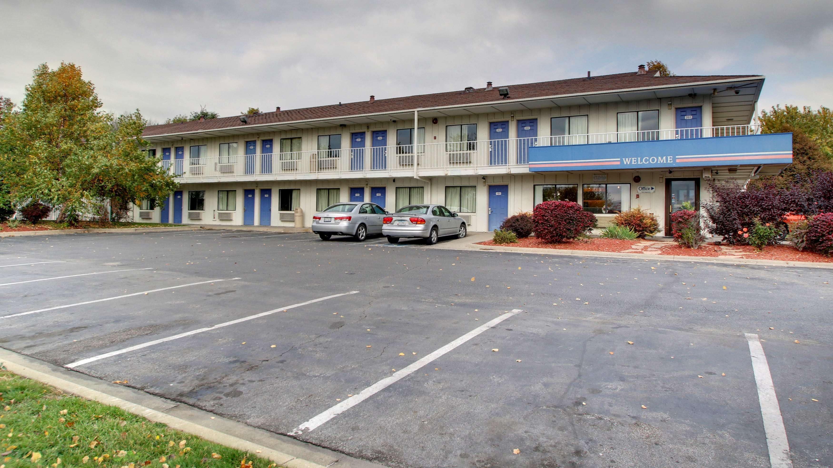 Un motel con dos coches estacionados en la entrada, un cartel de 'WELCOME' sobre la puerta y un pequeño aparcamiento.