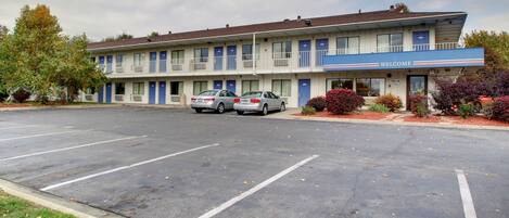 Un motel con dos coches estacionados en la entrada, un cartel de 'WELCOME' sobre la puerta y un pequeño aparcamiento.
