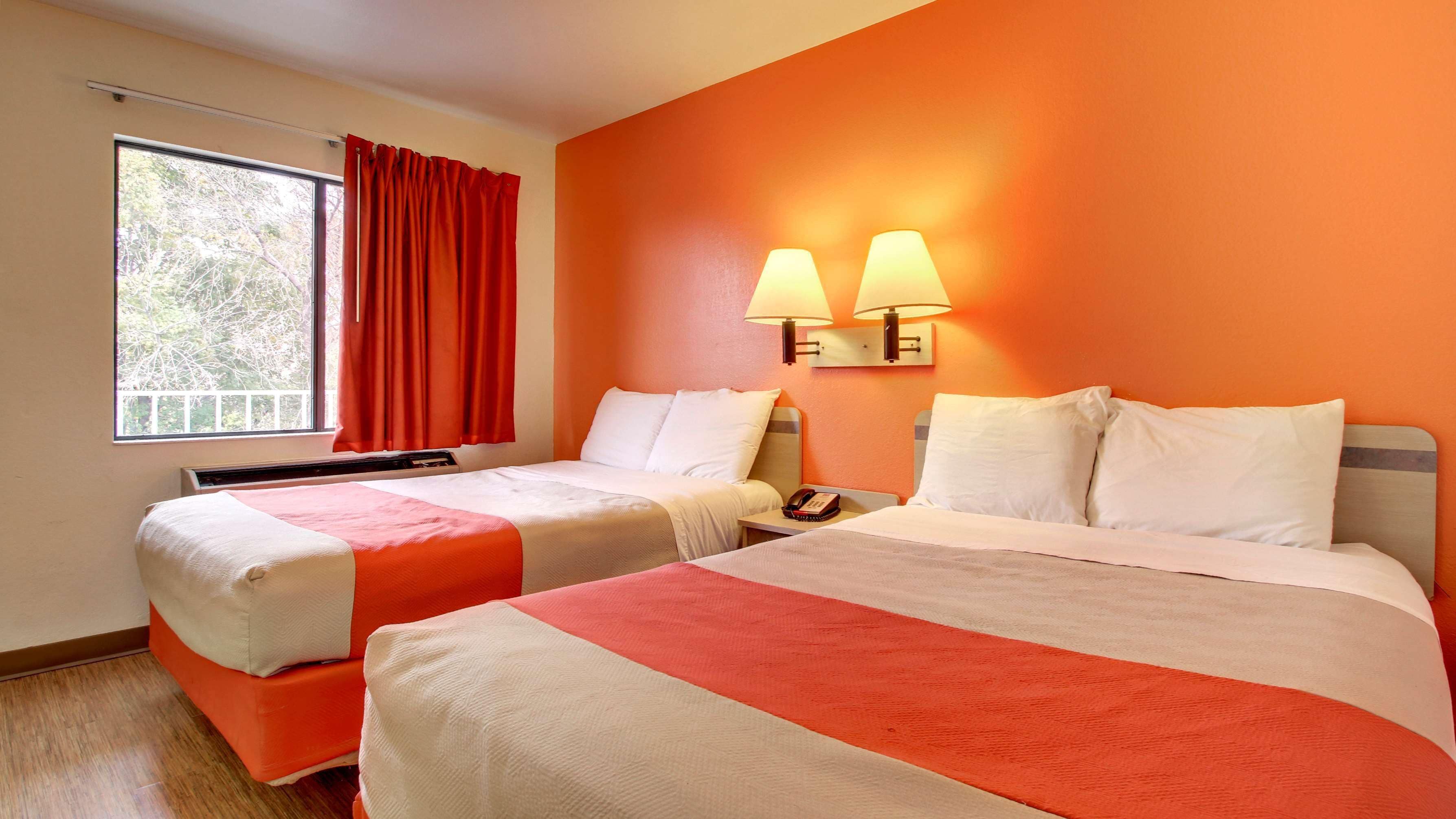 A hotel room with two beds, a window with red curtains, and two wall-mounted lamps.
