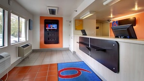 A clean, modern hotel lobby with a reception desk, a television screen, and a sign encouraging guests to save more for travel. - Motel 6 Des Moines, IA - North (Des Moines)