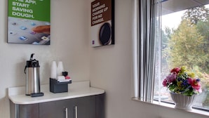 A coffee station with a coffee maker, cups, and a sign about saving dough. - Motel 6 Des Moines, IA - North (Des Moines)