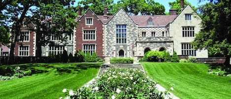 Un edificio grande e histórico con fachada simétrica, un patio central con un parterre floral y un césped bien cuidado.