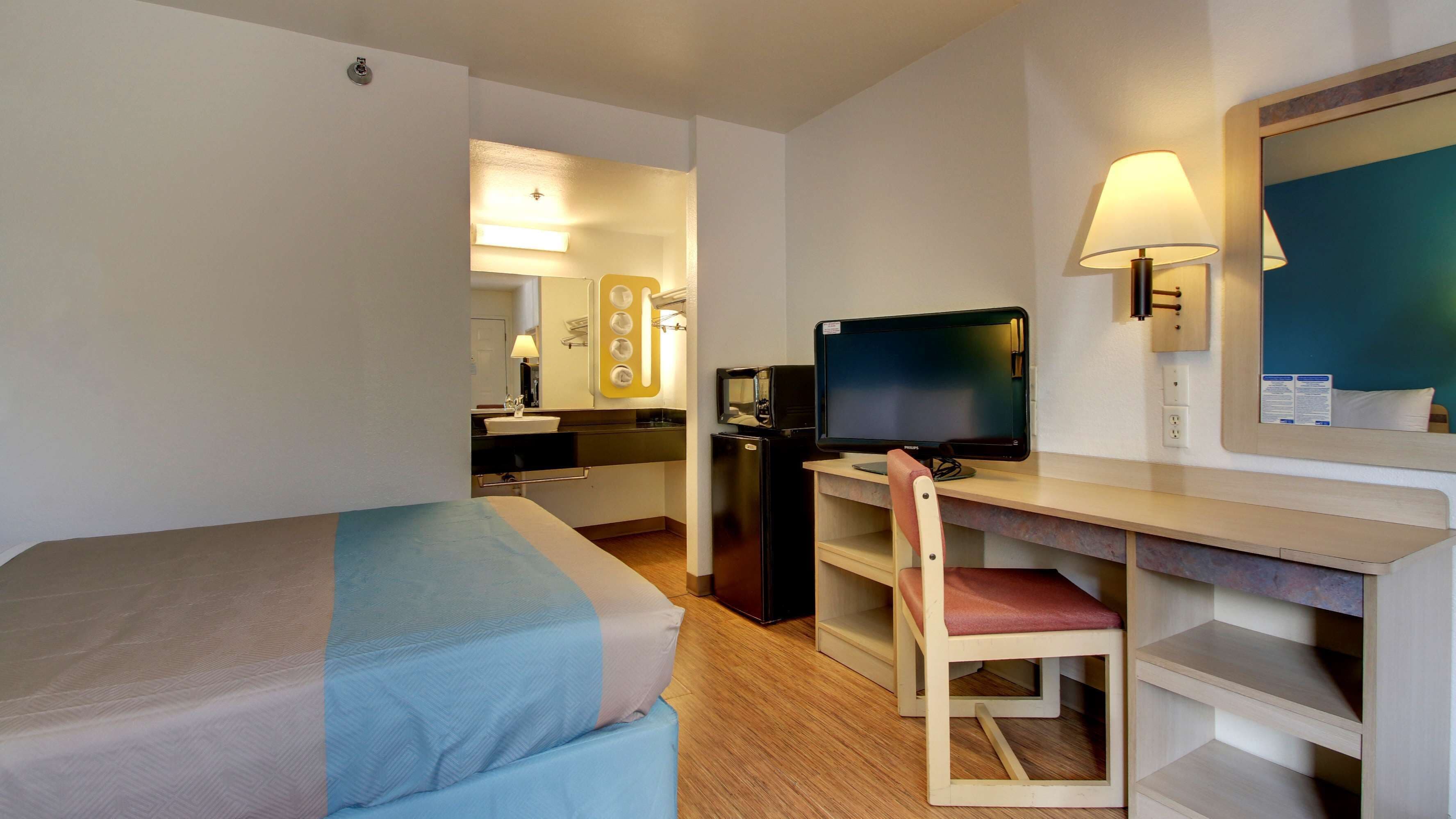 Une chambre d’hôtel comprenant un lit, un bureau avec un ordinateur, une chaise, un réfrigérateur, un miroir et une petite kitchenette.