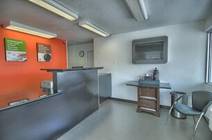 Una zona de recepción moderna con un mostrador gris, un pequeño armario de madera, un cesto de basura y una silla. Hay dos carteles en la pared: uno promociona una aplicación móvil y otro sobre servicios de carga.