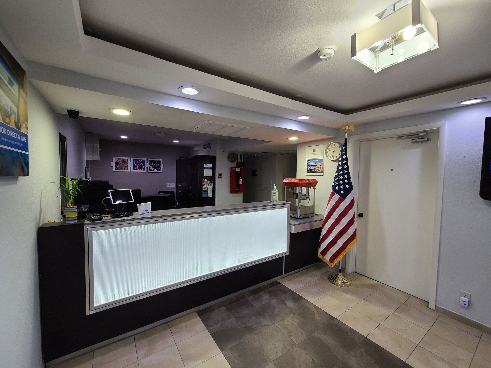 A reception area with a desk, computer, and an American flag. There are ceiling lights, a clock, and a fire extinguisher.
