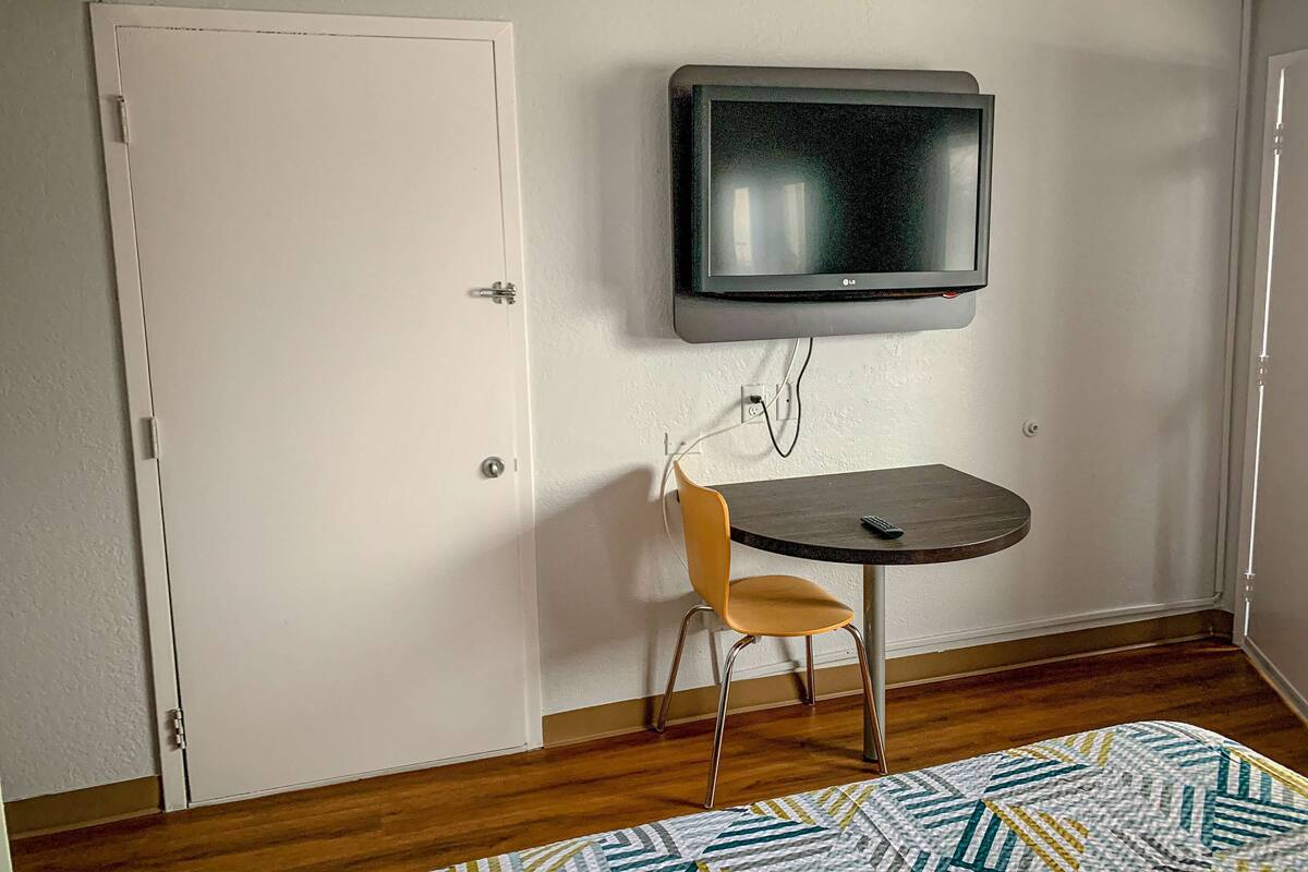 Ein Zimmer mit weißer Tür, einem an der Wand befestigten Fernseher, einem kleinen Tisch mit Stuhl und einem gemusterten Teppich auf dem Boden.