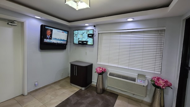 Un área de recepción con un televisor de pantalla plana, un mueble pequeño, una ventana con persianas y dos jarrones con flores.