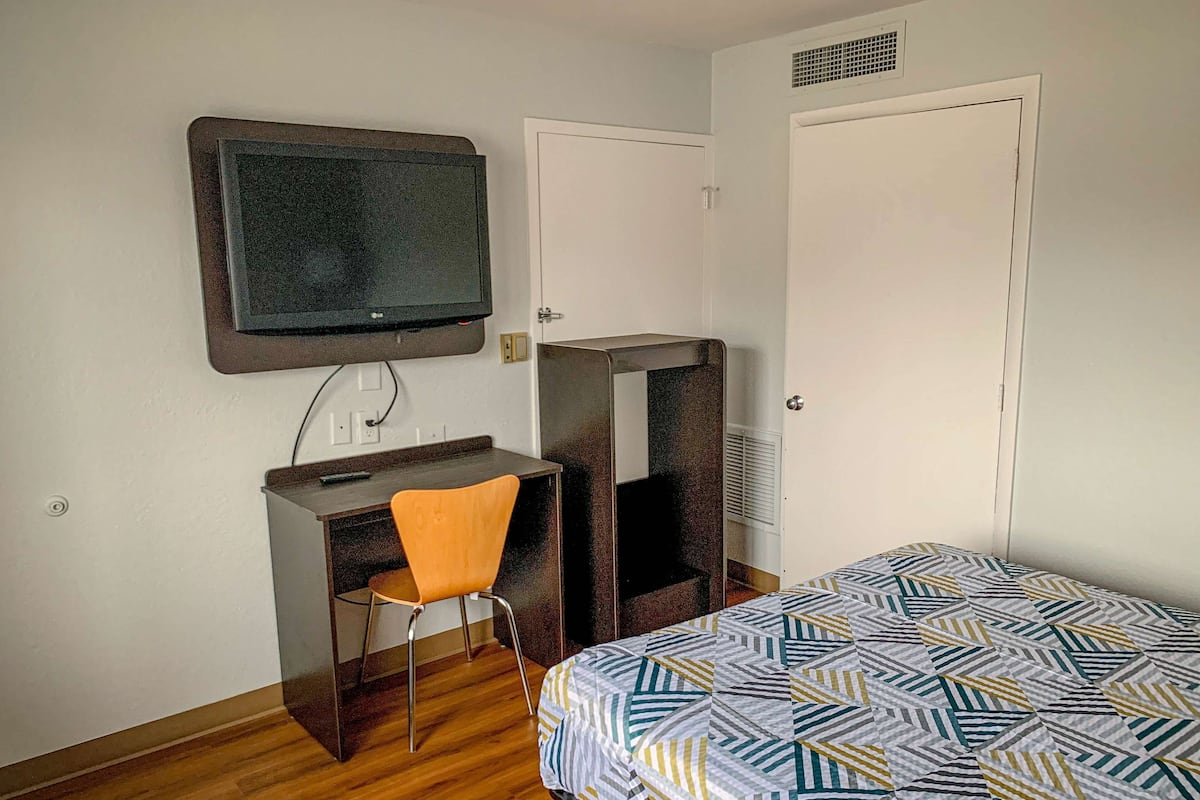 Ein Hotelzimmer mit Holzboden, einem Bett mit geometrischem Muster, einem Schreibtisch mit Stuhl, einem an der Wand befestigten Fernseher und einer Tür mit Spiegel.