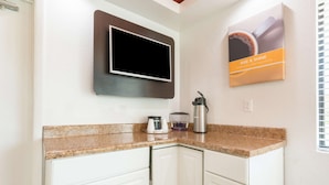 Una cocina con armarios blancos, encimera de granito, cafetera, hervidor y televisor montado en la pared.