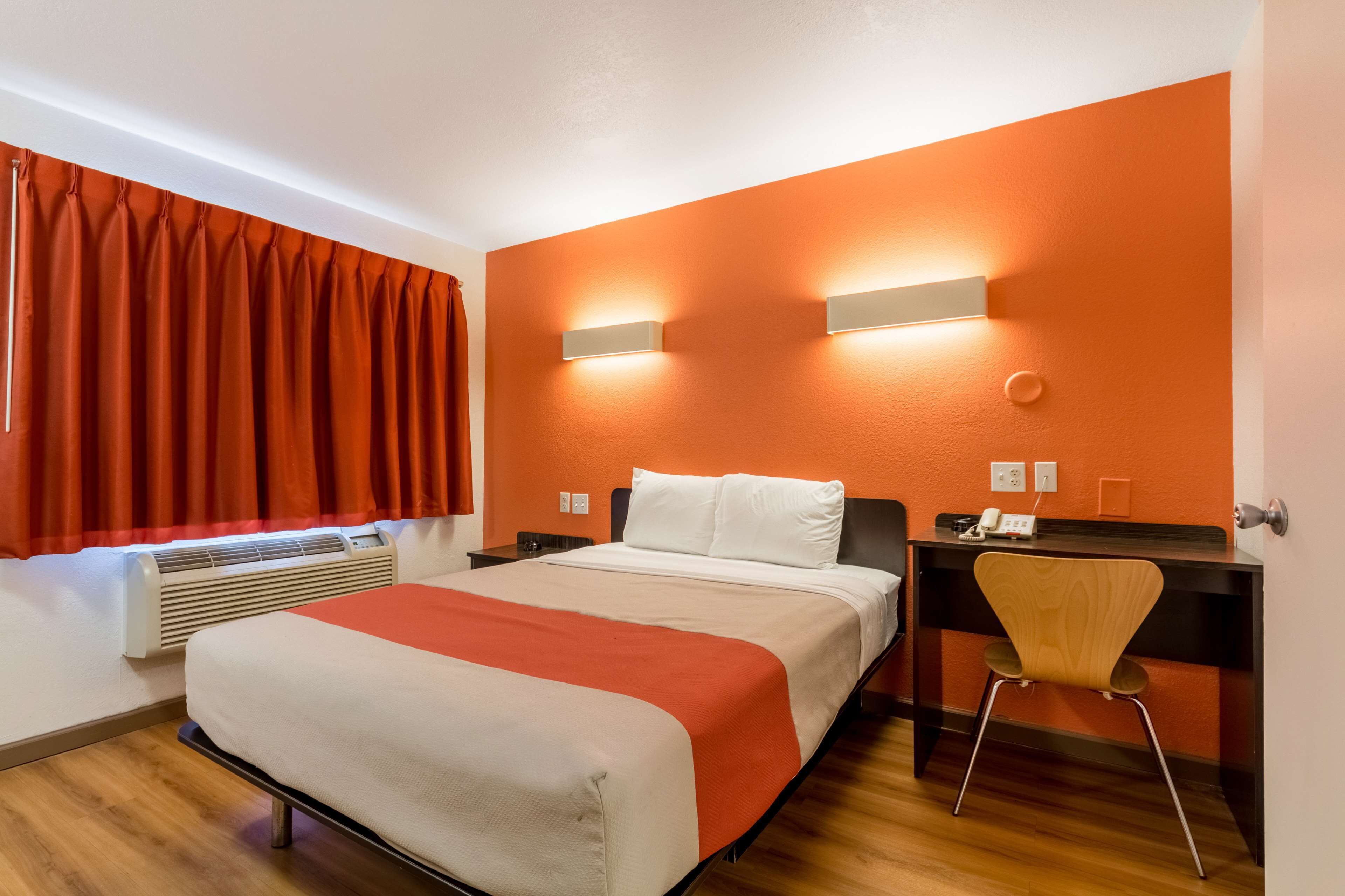 A hotel room with a bed, a desk, a chair, and a wall-mounted light fixture.