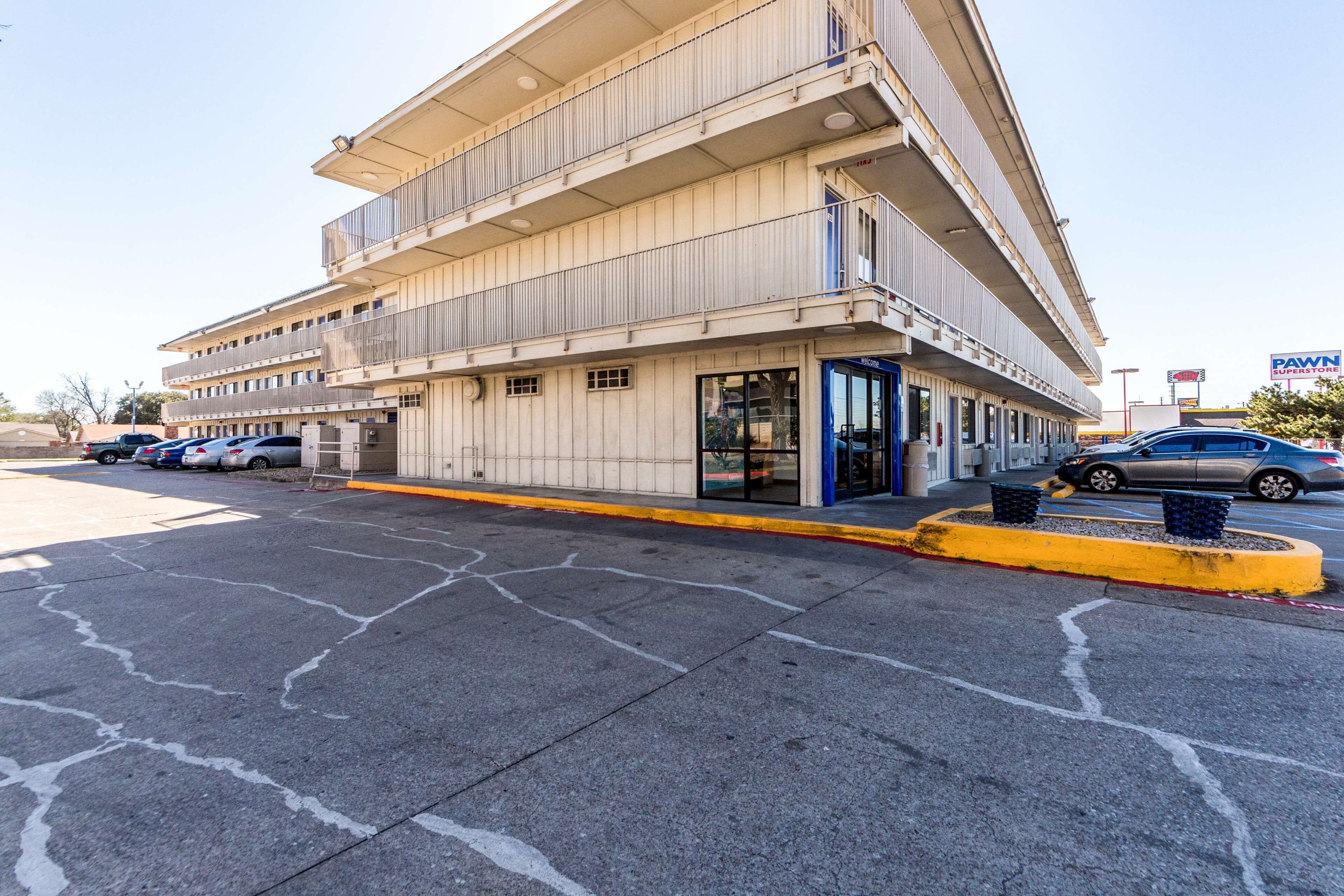 Um edifício de vários andares com fachada plana, entrada visível com portas de vidro e um estacionamento com vários carros.