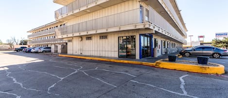 Um edifício de vários andares com fachada plana, entrada visível com portas de vidro e um estacionamento com vários carros.