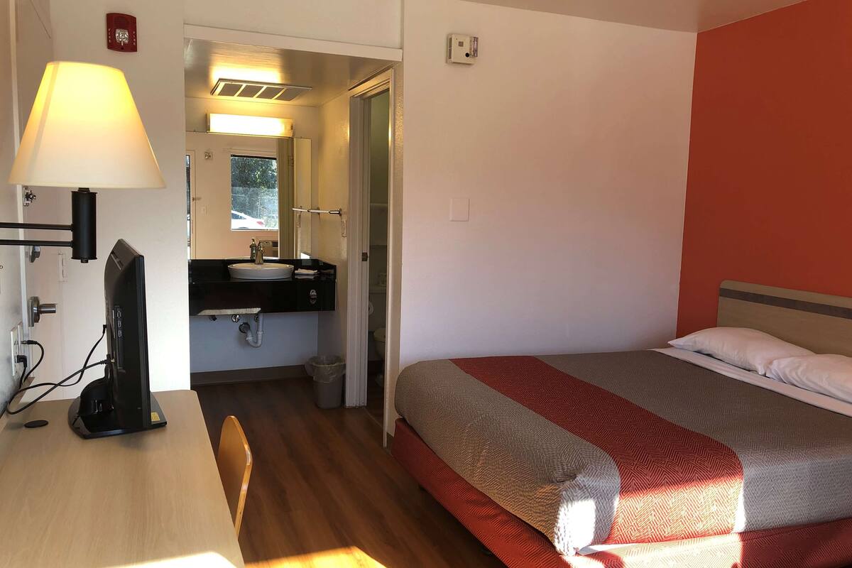 Ein Hotelzimmer mit Schreibtisch, Bett mit gemustertem Überwurf, Badezimmer mit Waschbecken und Spiegel sowie einer roten Wand.