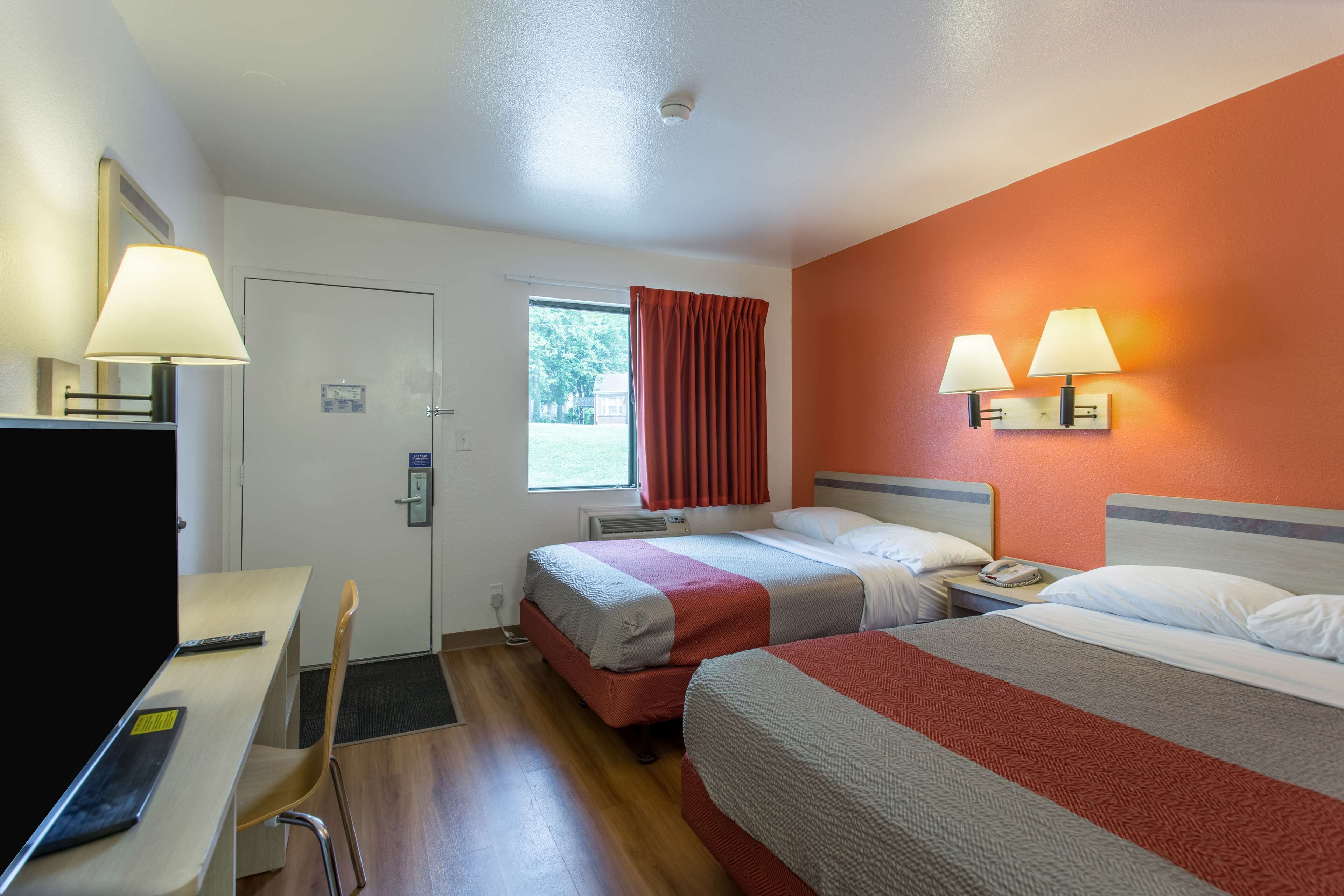 Ett hotellrum med två sängar, ett skrivbord och en stol. Det finns ett fönster med gardiner, en vägglampa och en dörr med en skylt.