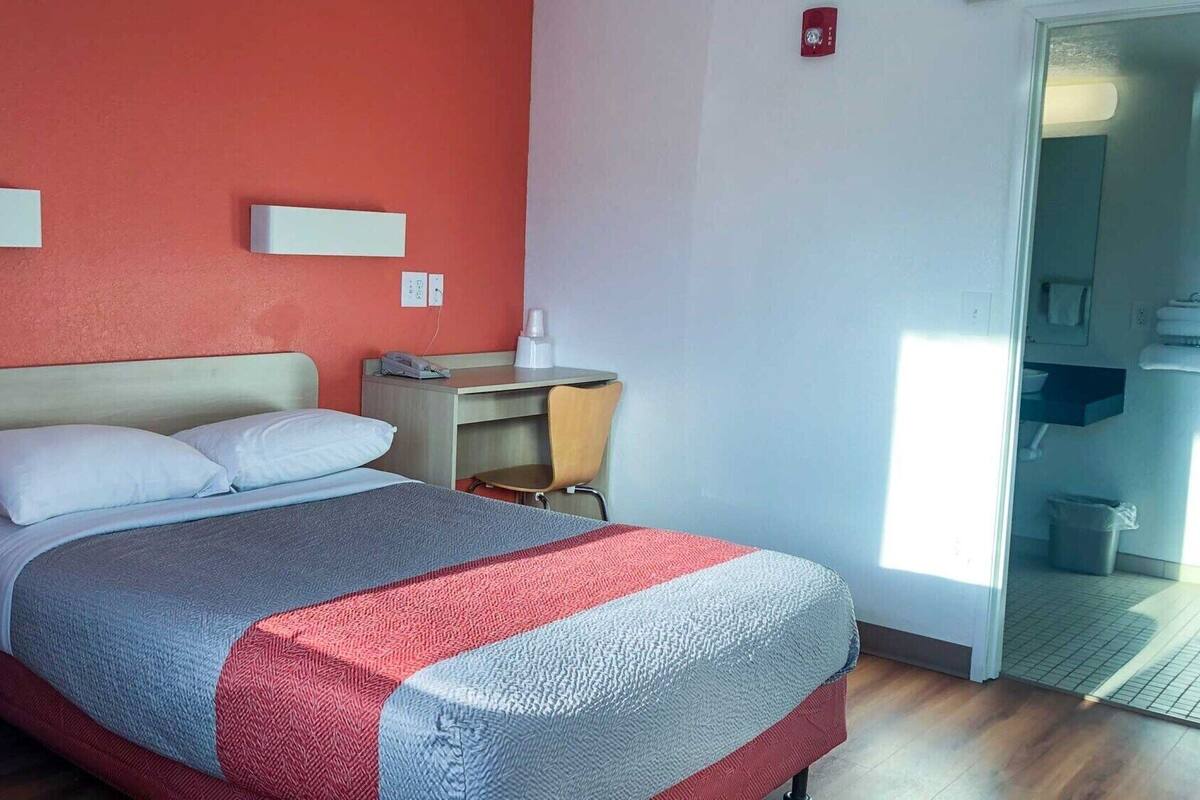 Ein Hotelzimmer mit Bett, Nachttisch und Schreibtisch mit Stuhl. Ein Fenster sorgt für natürliches Licht und bietet einen Blick ins Freie.