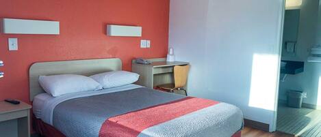 Quarto de hotel com cama, mesa de cabeceira e escrivaninha com cadeira. Há uma janela que oferece luz natural e vista para o exterior.