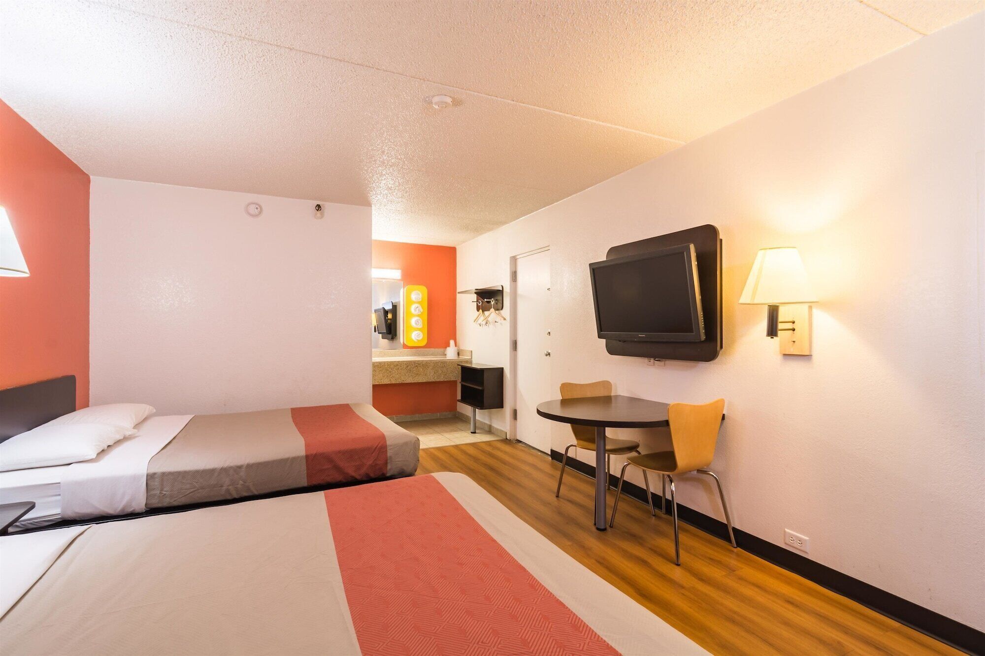 A hotel room with two beds, a TV, a small table with a chair, and a bathroom visible in the background.
