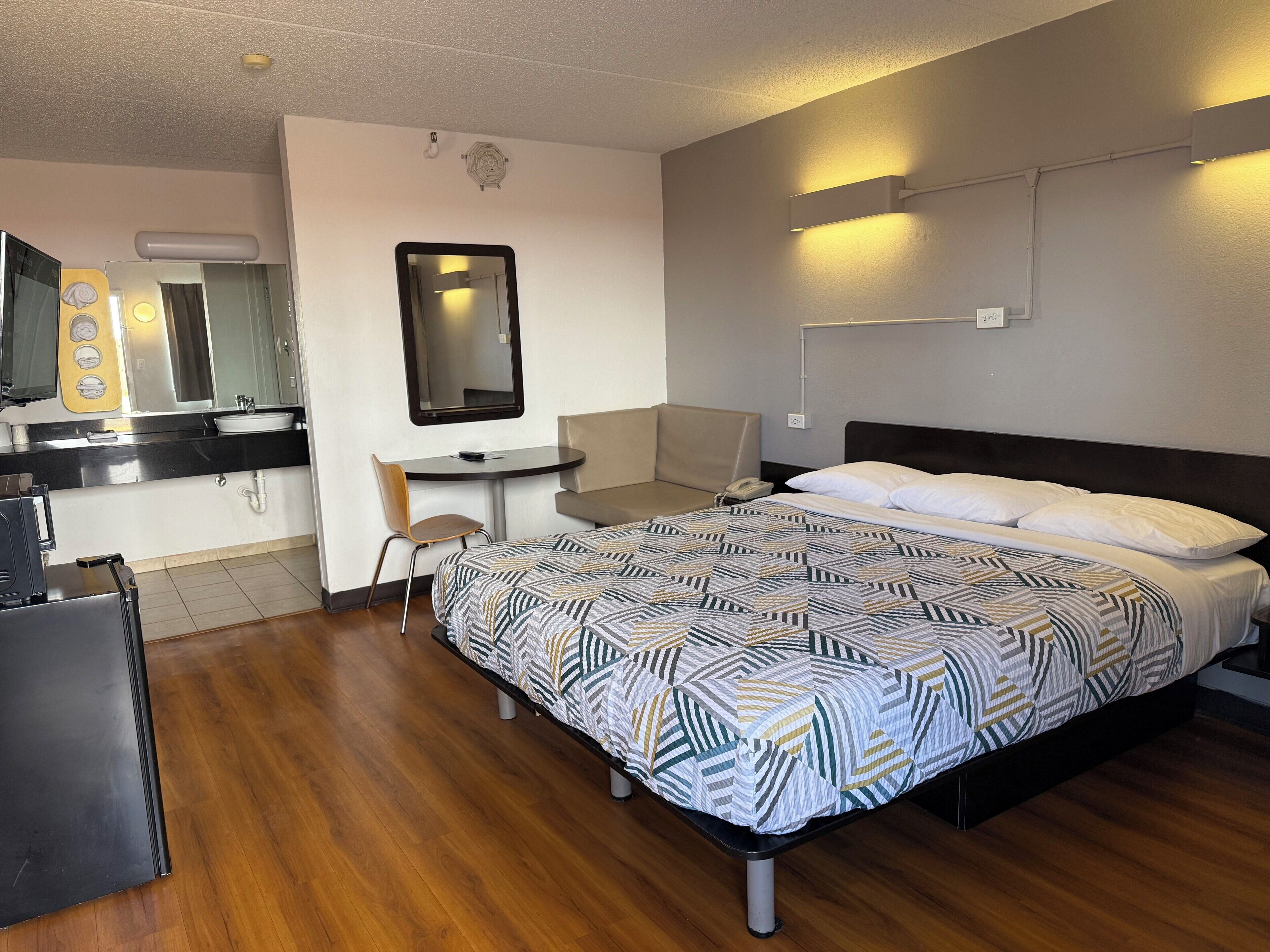 Habitación de hotel con cama, escritorio, silla, espejo, lavabo y un refrigerador pequeño.