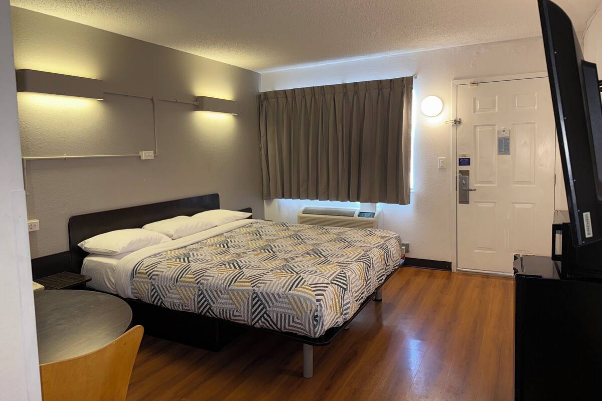 A hotel room with a bed, a desk, a chair, a TV, a window with curtains, and a door.