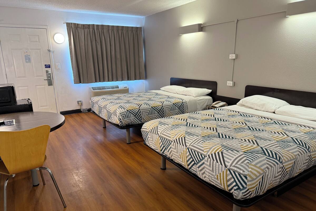 A hotel room with two beds, a desk, a chair, a window with curtains, and a wall-mounted air conditioner.