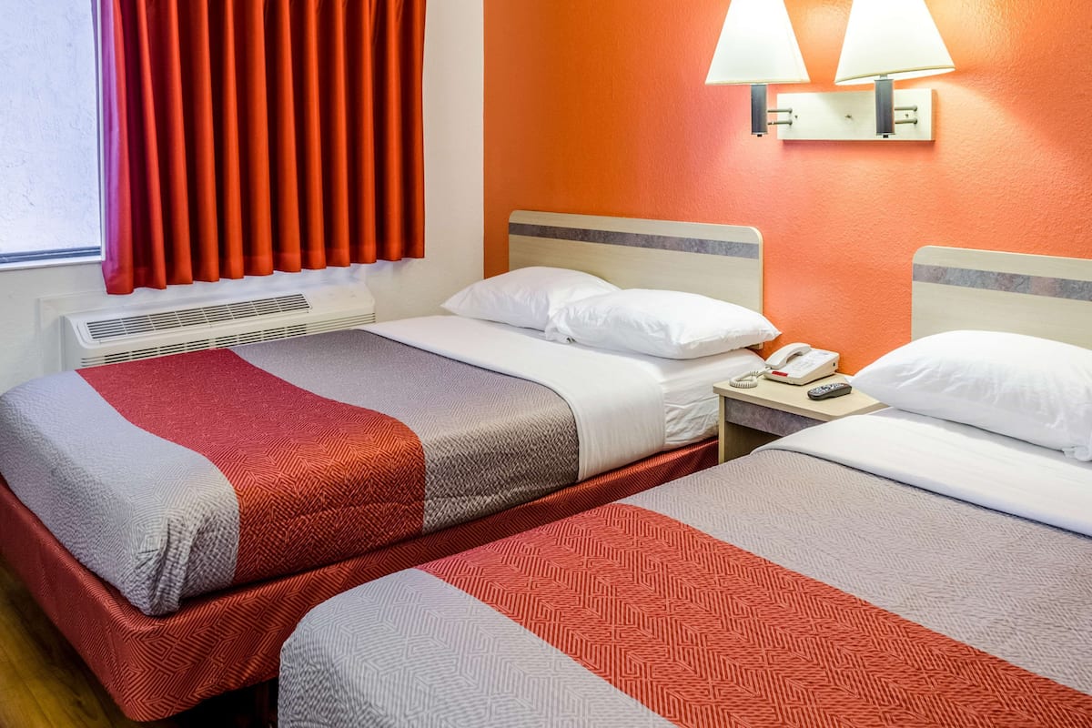 Twee eenpersoonsbedden met rood en grijs beddengoed, een witte nachtkastje met een telefoon en een lamp, en een airconditioning.