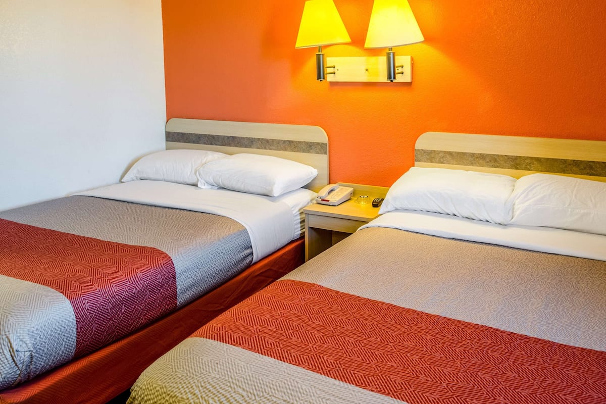 Een hotelkamer met twee bedden, elk met een bedovertrek met een patroon en witte kussens. Daartussen staat een nachtkastje met een telefoon en een lamp. De muren zijn oranje geschilderd.